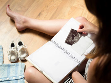 Woman Sitting on Floor Looking at Baby Sonograph in Notebook Surrounded by Other Babby Keepsakes - KeepsakeMom
