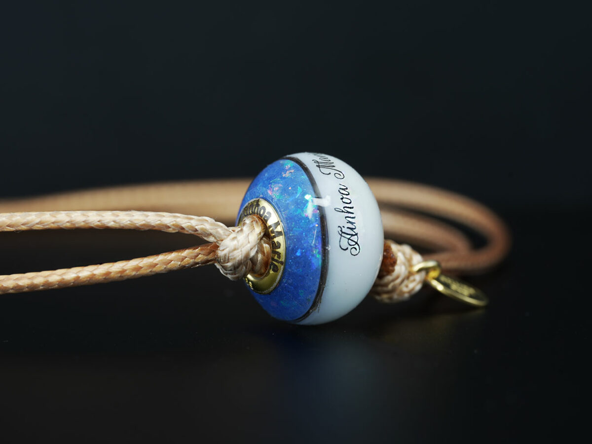 breastmilk one bead bracelet with birth month colors of sapphire blue, lock of hair and names on the breastmilk part of the bead on a waxed light brown cord on black background from KeepsakeMom