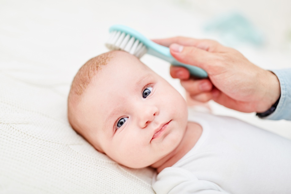 Baby with cradle cap lying on bed while parent brushes hair