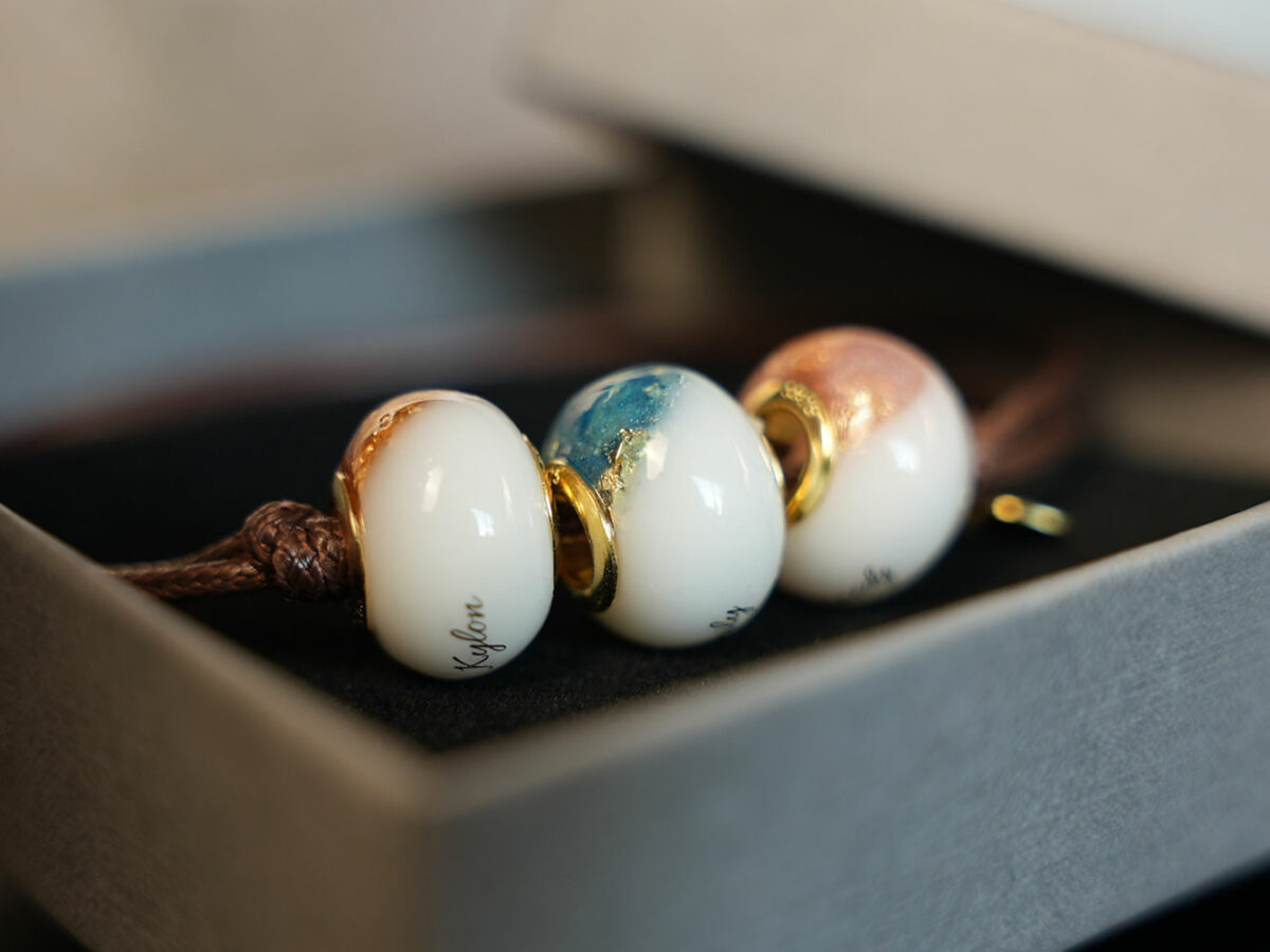 three breastmilk beads with birth month colors pink, orange and blue with gold flakes at the top and children names in the milk engraved with gold cores on a brown waxed band bracelet in a box from KeepsakeMom