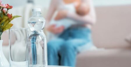 Close-up view of glass, bottle of water, flowers in vase and mother breastfeeding baby behind at home