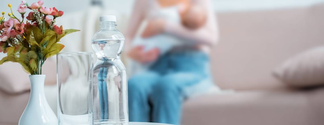 Close-up view of glass, bottle of water, flowers in vase and mother breastfeeding baby behind at home