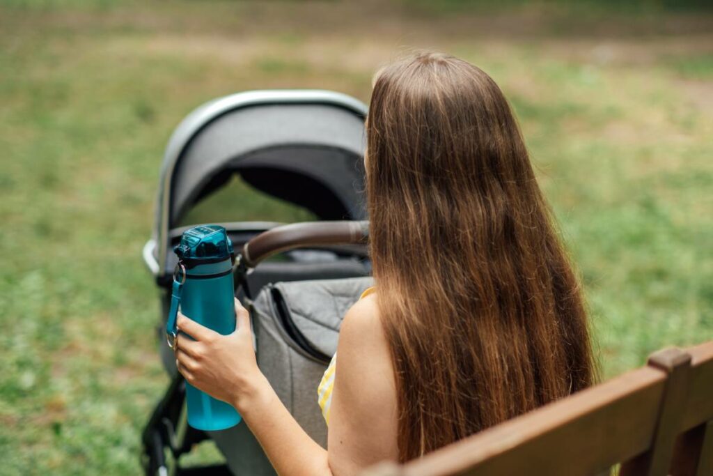 Young mother with a water bottle in hand, pushing newborn baby in stroller 