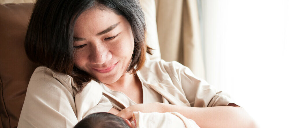 Mother breastfeeding her newborn baby beside window in cross cradle position.