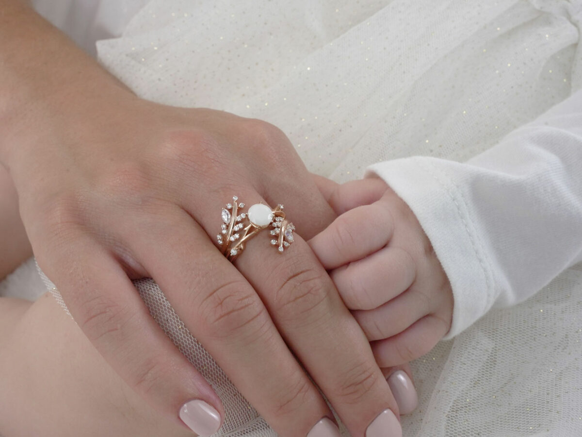 breastmilk jewelry two rings set in gold plated rose gold over sterling silver with central small oval milk stone and leaf branches fanning out on the outer ring from KeepsakeMom on mother hand holding baby hand