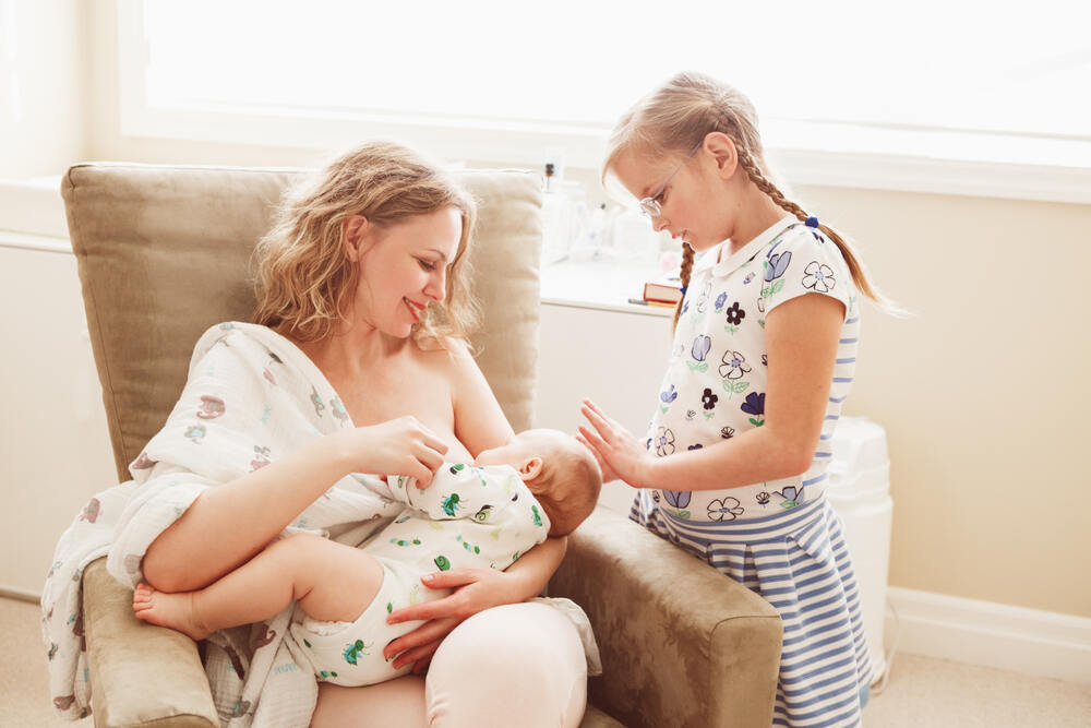 Woman breastfeeding baby in chair with older sibling by side