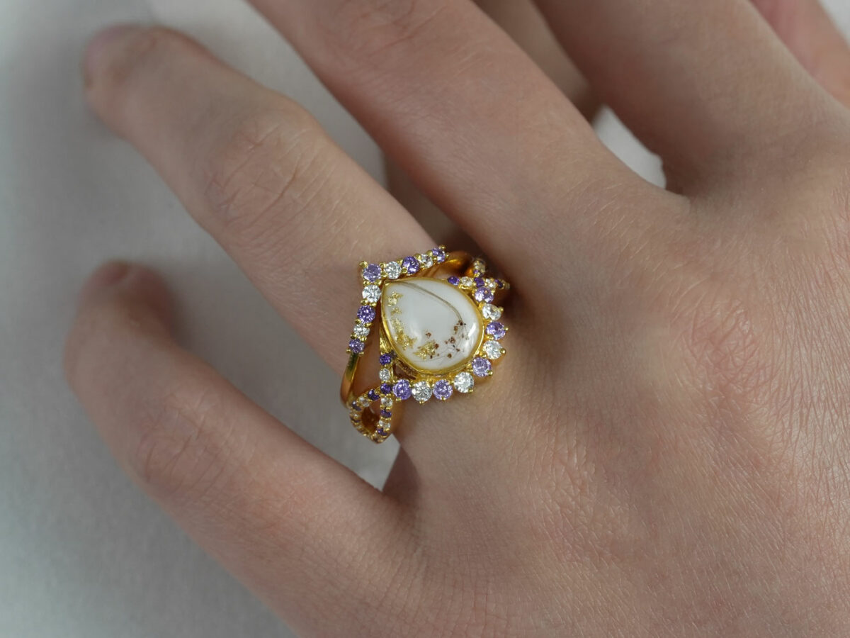 breast milk ring with birth month crystals around teardrop shaped milk stone with hair and placenta and yellow gold plating from keepsakemom on a model hand
