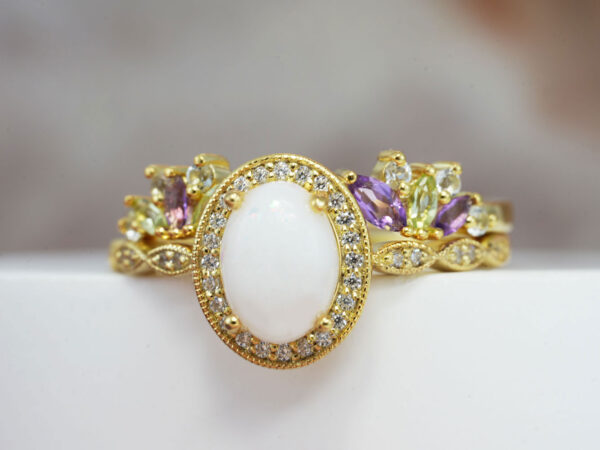 breastmilk ring set of two yellow gold rings with fine band and clear crystals around with oval breastmilk stone and prongs holding the stone and add -on ring on top with purple and peridot birth month stones in wings shape front view on a large stone from KeepsakeMom