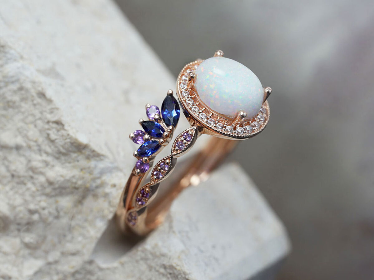 breastmilk ring set of two rose gold rings with fine band and purple crystals around with oval breastmilk stone and prongs holding the stone and add -on ring on top with sapphire blue birth month stones in wings shape side view on a large stone from KeepsakeMom