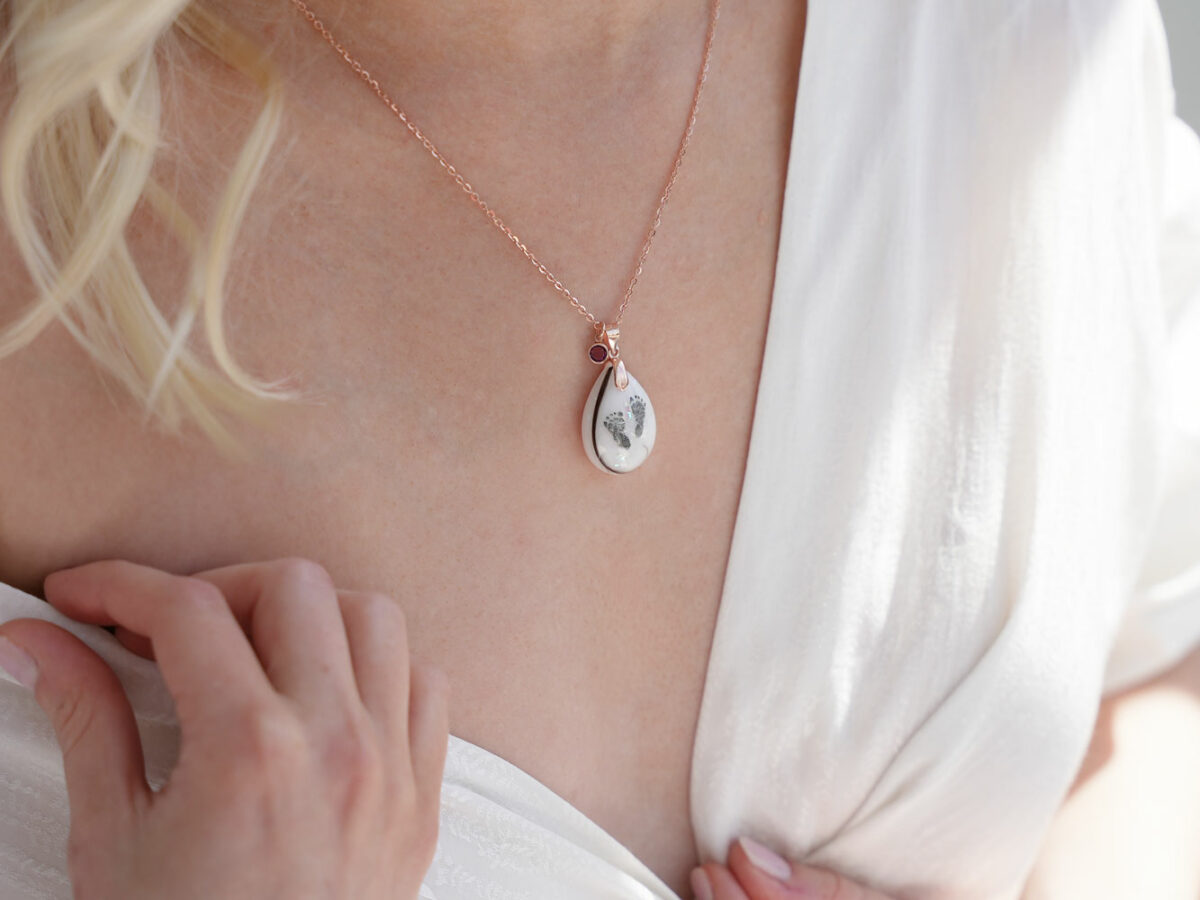 breastmilk teardrop necklace with baby foot prints lock of hair on a rose gold chain with garnet crystal on a model from KeepsakeMom