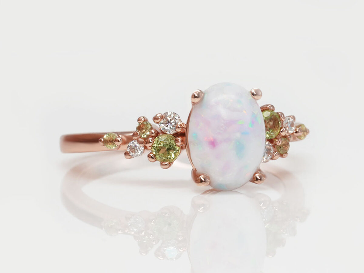 breast milk ring on a white background in rose gold and green peridot crystals on a side and central oval milk stone with opals blue and pink from keepsakemom