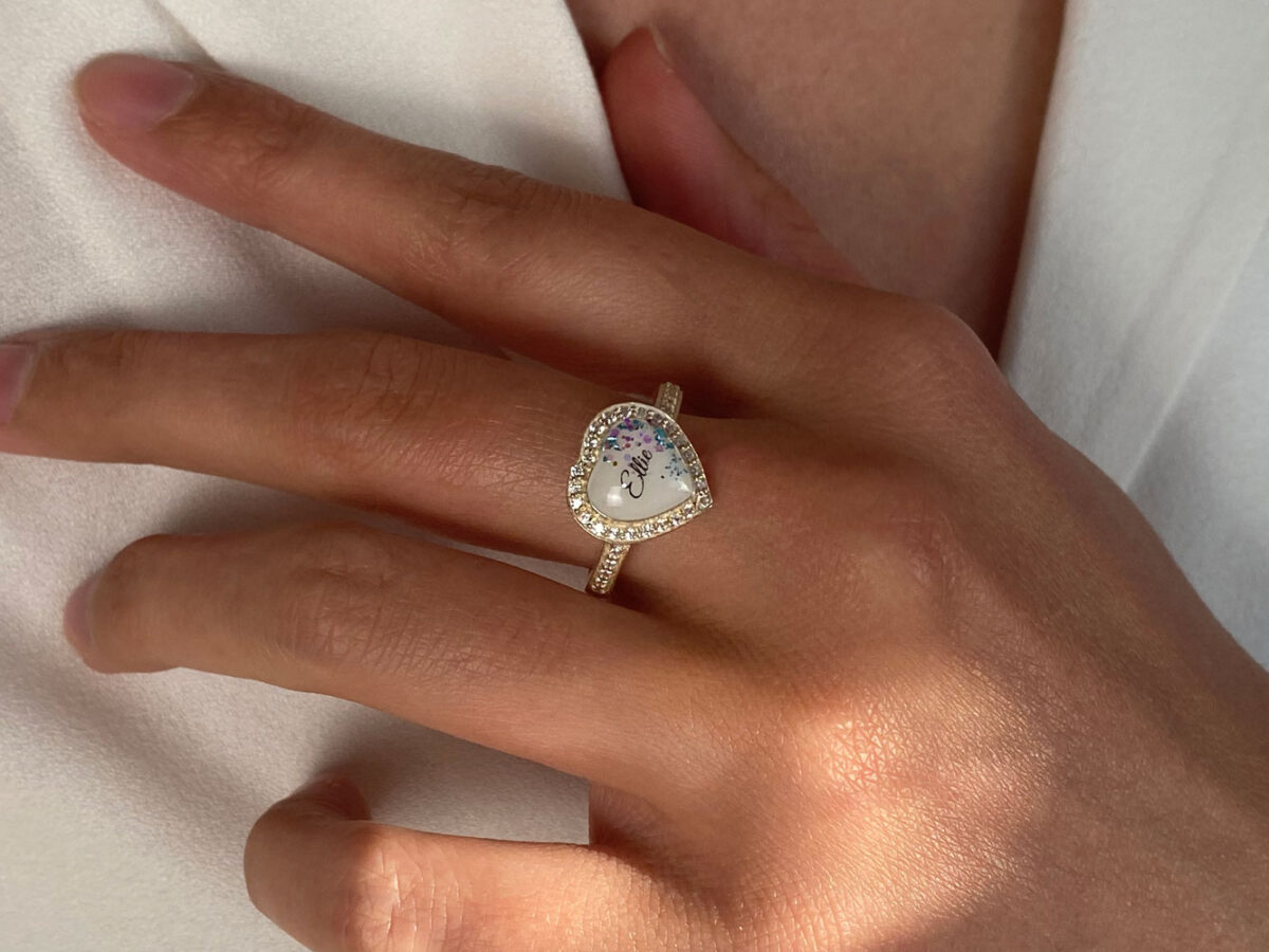 breastmilk ring in heart shape with halo clear crystals, name and colored flakes inside breastmilk stone for diy kit keepsakemom on a model hand