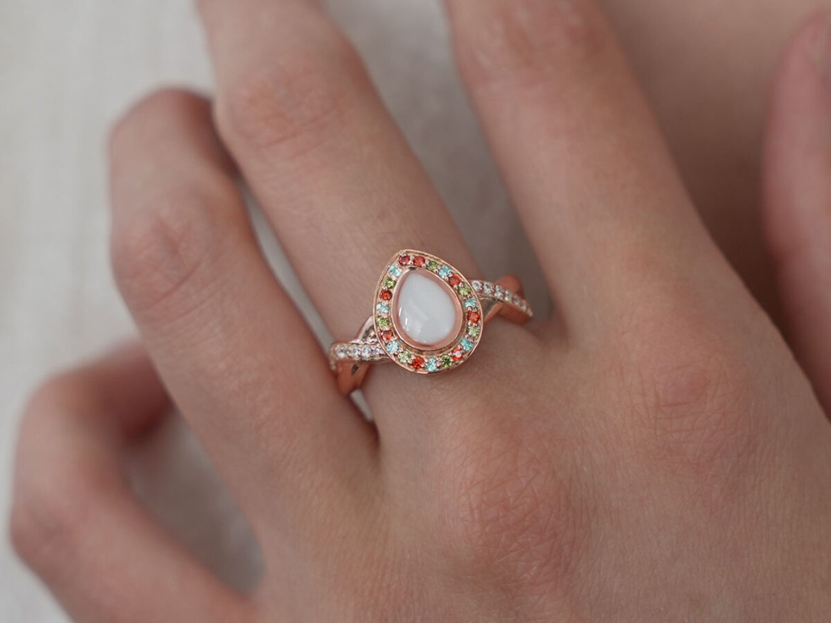 breastmilk ring with heart shaped central breastmilk stone with ruby red and green and blue gems as halo around the stone and around the ring band alternating from keepsakemom in rose gold setting on models hand