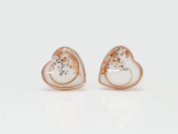 memorial jewelry a pair of rose gold heart shaped earrings with ashes, rose gold flakes and locks of hair layered on white backdrop of breast milk filled cups from keepsakemom