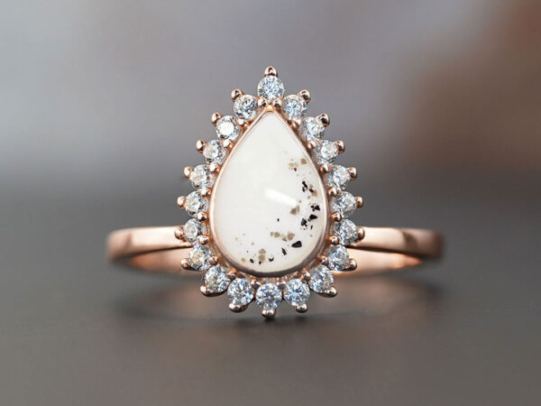 memorial cremation teardrop shaped ring with ashes on a dark brown background, adorned with clear crystals in rose gold filled with breastmilk and ashes and lock of hair from keepsakemom